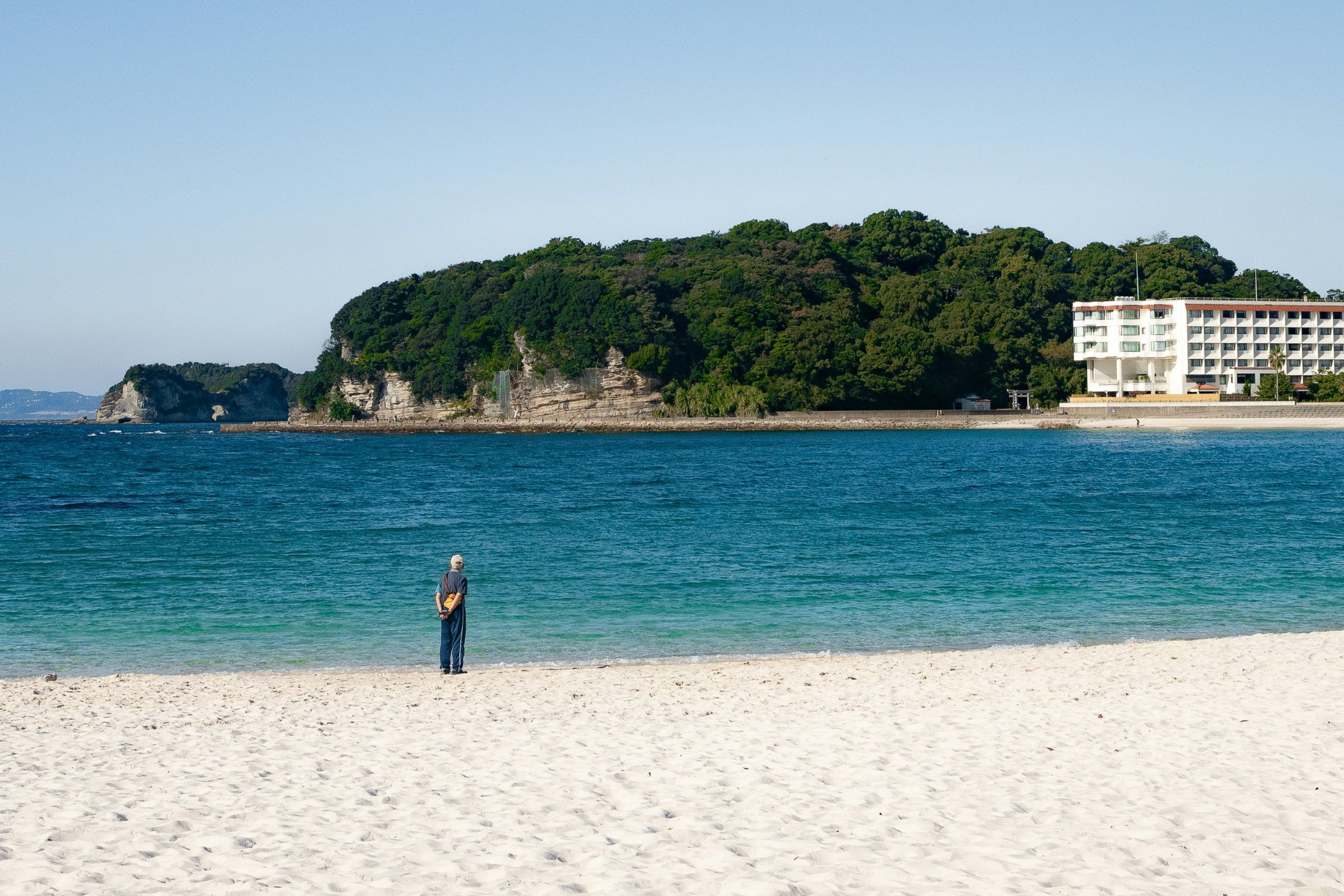 I forgrunden ses en bred, hvid sandstrand. I vandkanten står en mand og kigger ud over det turkisblå vand. Billedet er fra Shirahama Beach i Japan. 