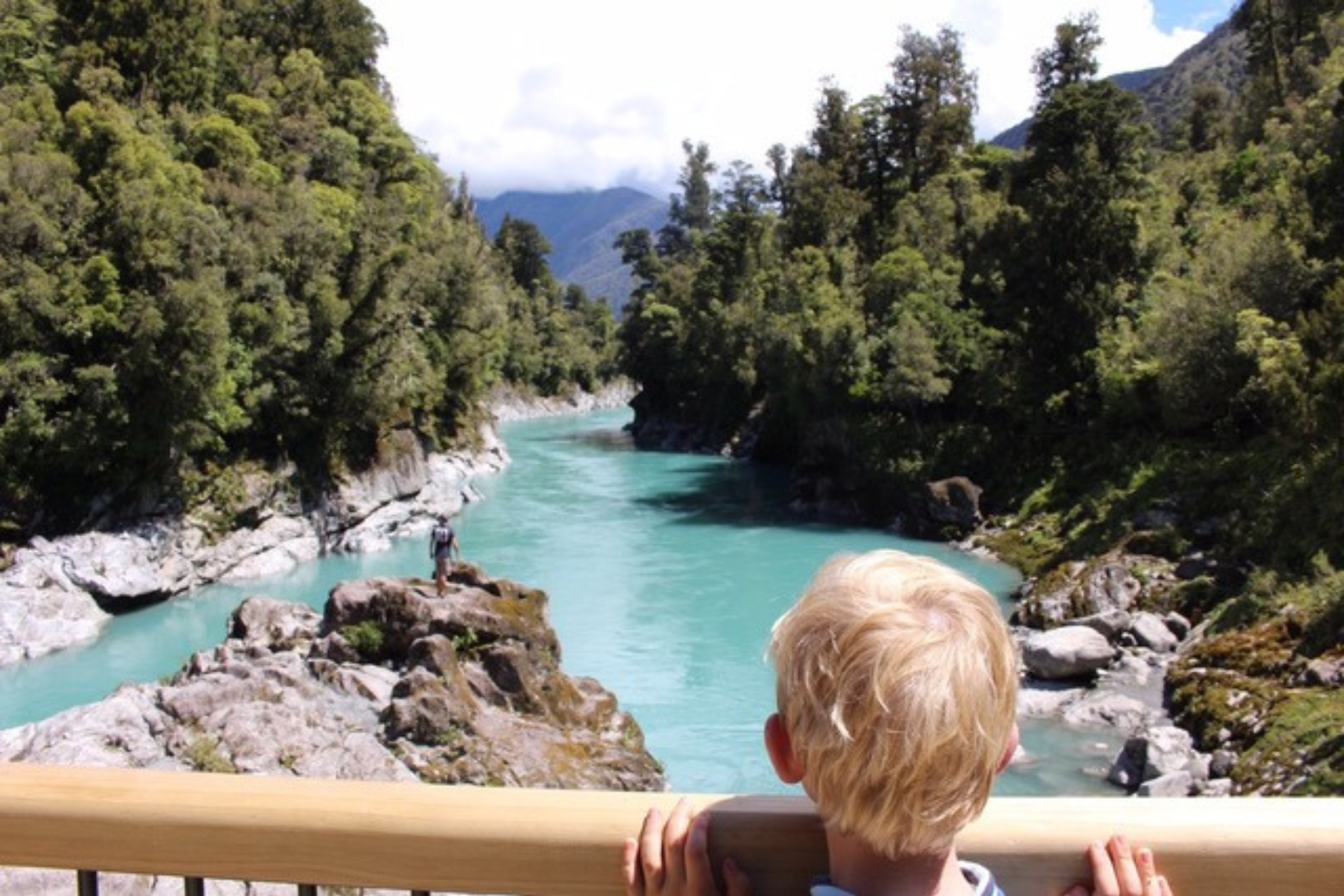 En lyshåret dreng står og kigger ud over en turkisgrøn flod. 