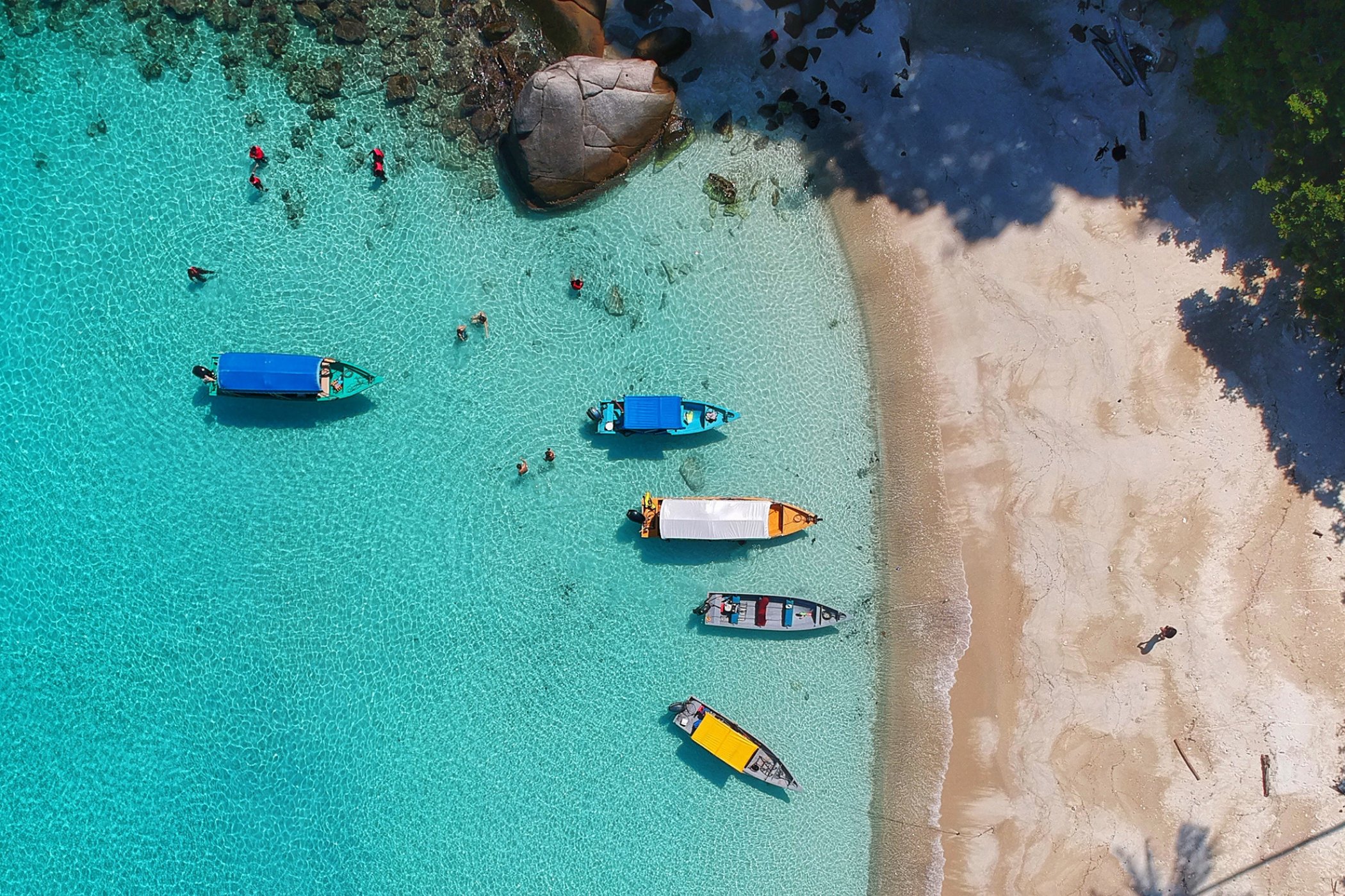 Turkisblåt hav og sandstrand set oppefra - der er fem både i vandet. 