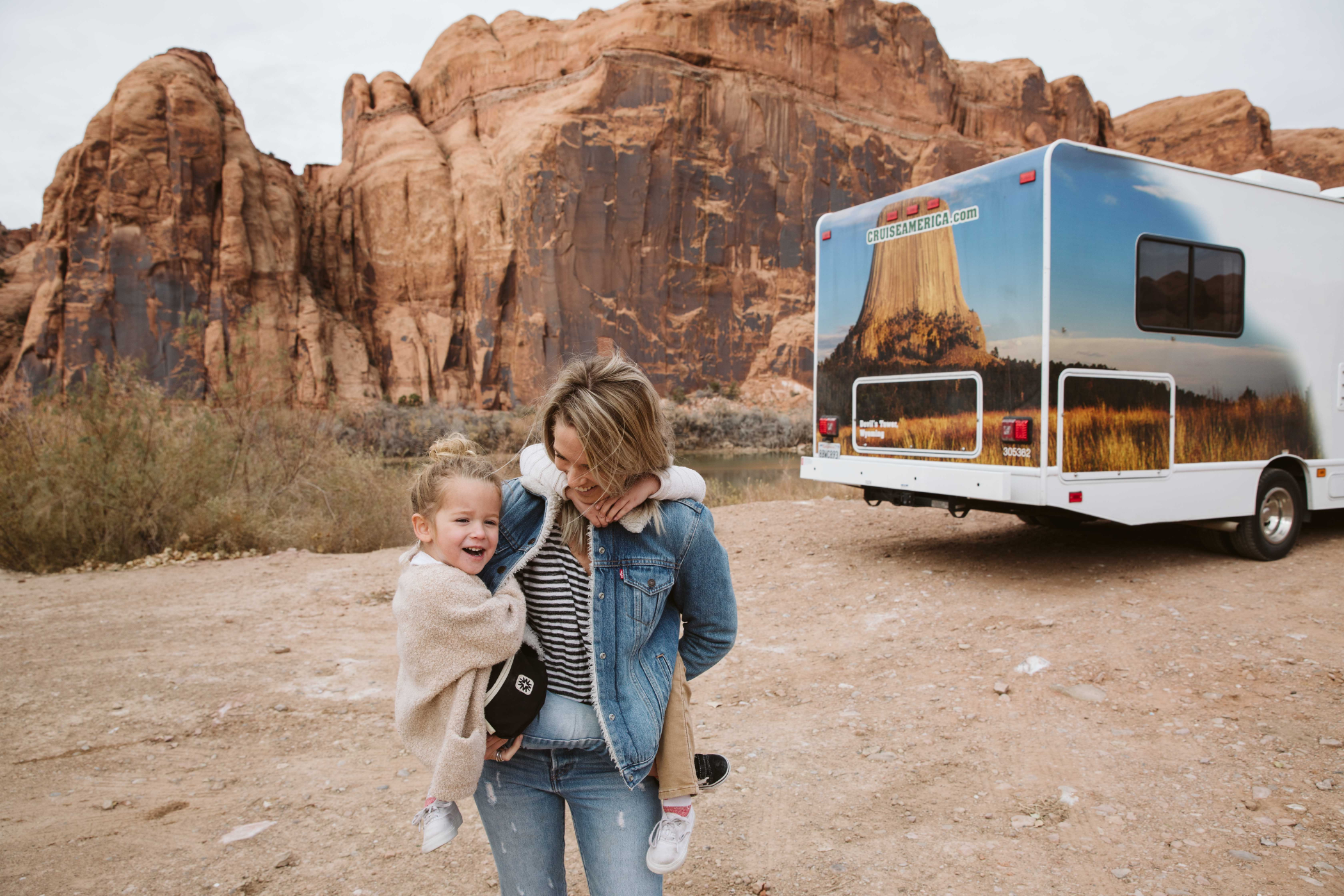 En kvinde bærer to mindre børn og står foran en autocamper og klipper. 
